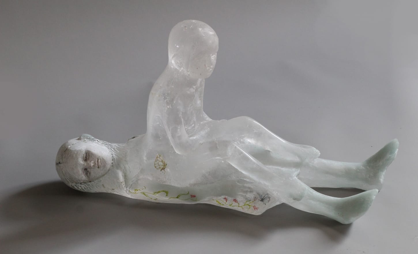 a glass sculpture of a child laying on the ground and another seeming to sit up out of her, like a spirit