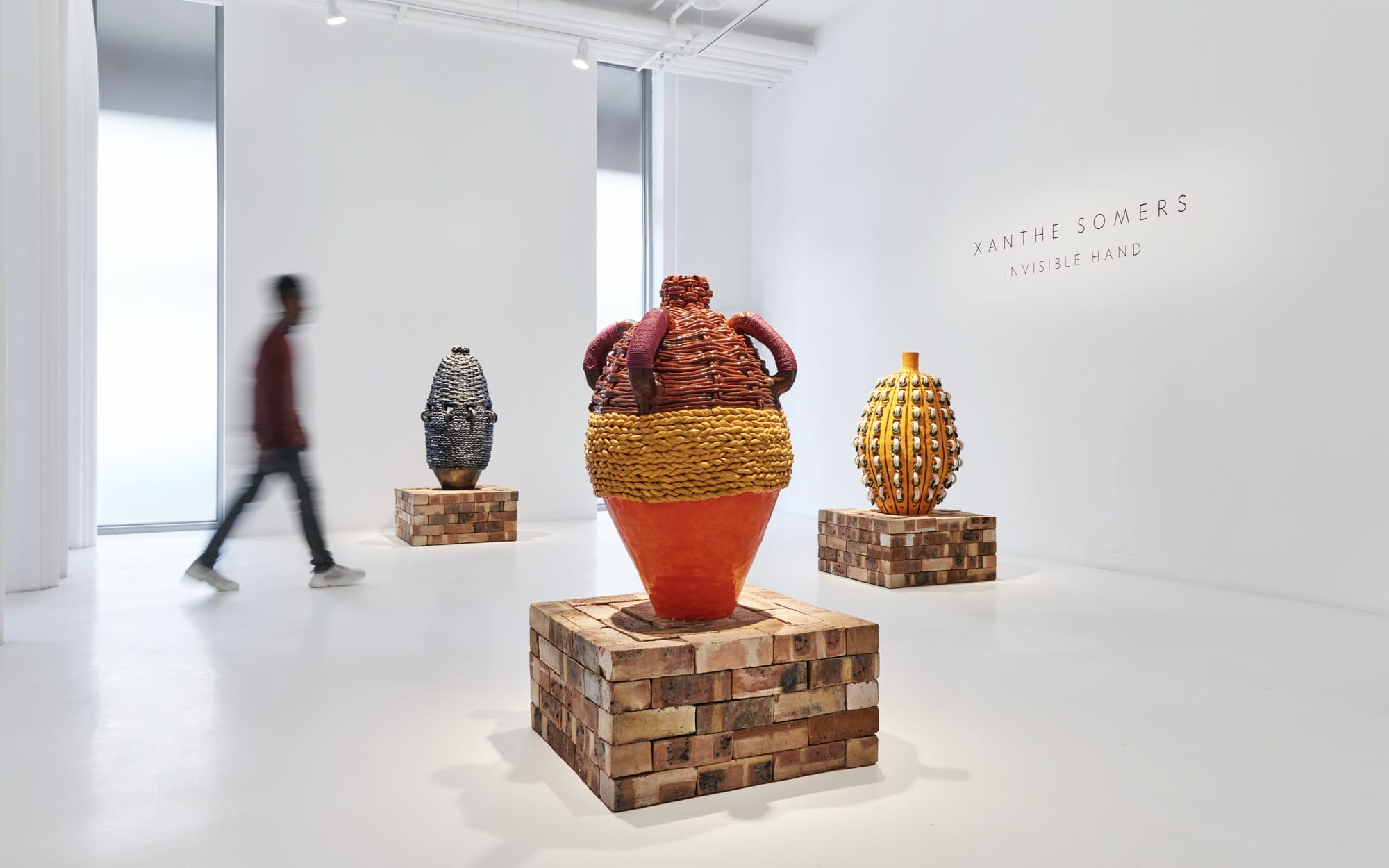 a person walks through a white-wall gallery space filled with colorful, large-scale ceramic vessels